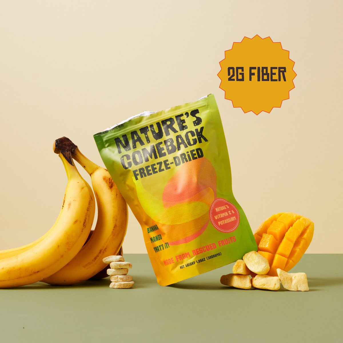 A bag of Nature's Comeback Freeze-Dried fruit snacks is displayed on a green surface with bananas, mango slices, and a few dried snack pieces around it. The packaging is yellow and green with a red circular badge, and an orange starburst graphic in the top right says '2G FIBER'.
