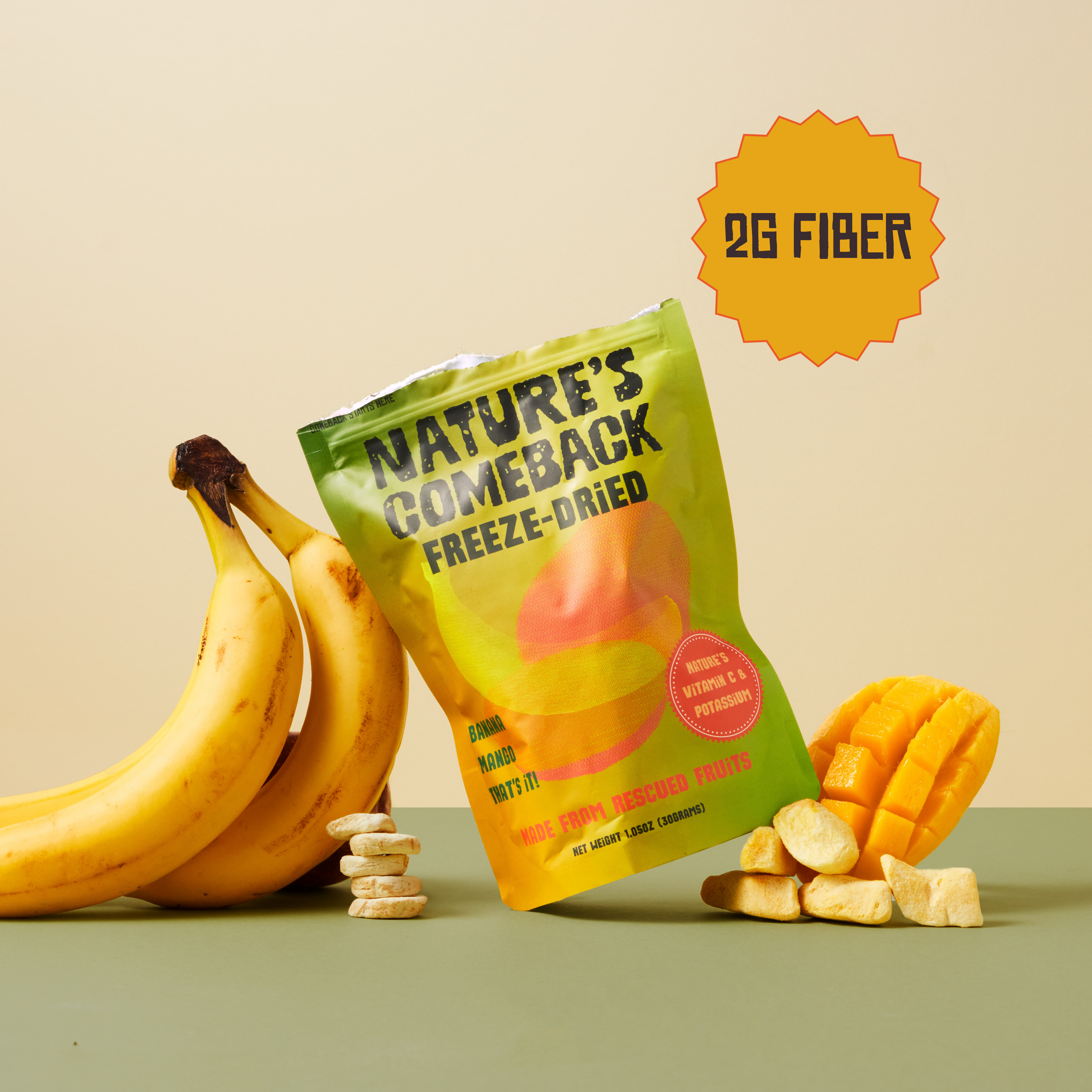 A bag of Nature's Comeback Freeze-Dried fruit snacks is displayed on a green surface with bananas, mango slices, and a few dried snack pieces around it. The packaging is yellow and green with a red circular badge, and an orange starburst graphic in the top right says '2G FIBER'.